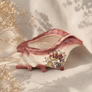 Può includere: Una fioriera in ceramica a forma di conchiglia nei toni del rosa e del bianco. La fioriera presenta un motivo floreale con bacche rosse e viola e foglie verdi. La fioriera ha un esterno strutturato.