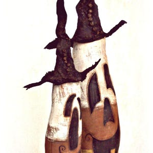 May include: Two tall, cylindrical figures with white, orange, and yellow color-blocked bodies featuring carved-out, dark-colored facial expressions. Each figure wears a tall, pointed, dark-colored hat adorned with a vertical row of small, round buttons.