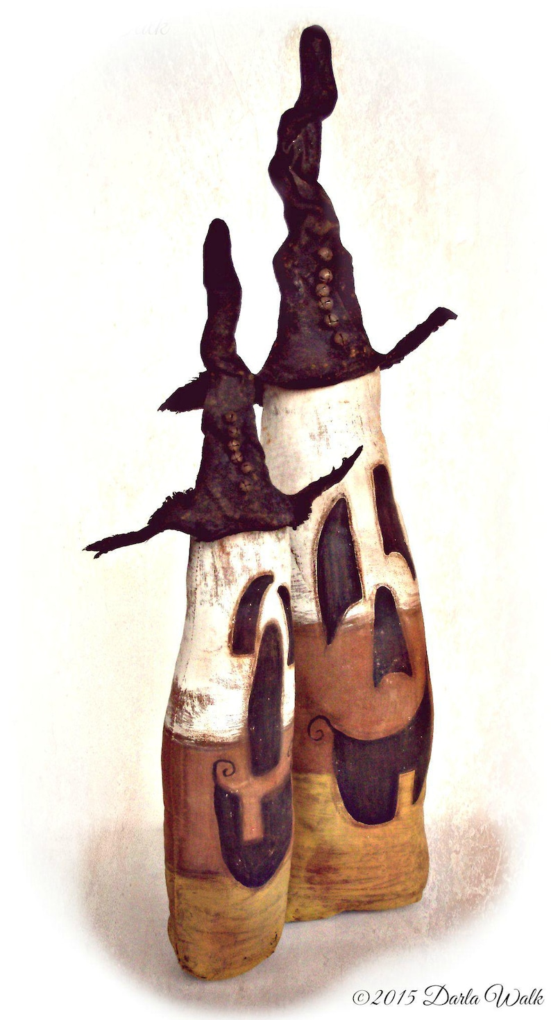 May include: Two tall, cylindrical figures with white, orange, and yellow color-blocked bodies featuring carved-out, dark-colored facial expressions. Each figure wears a tall, pointed, dark-colored hat adorned with a vertical row of small, round buttons.