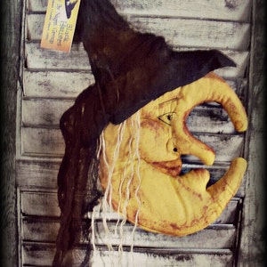 Puede incluir: Una luna amarilla en forma de creciente con una cara de bruja y cabello largo y blanco. La luna est&aacute; colgada en una contraventana de madera blanca con un sombrero de bruja negro en la parte superior. El sombrero tiene una etiqueta que dice "Black Hatters Fly By Night Group Salem Coven Since 1998".