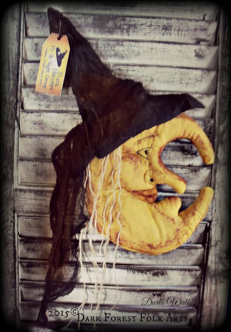 Puede incluir: Una luna amarilla en forma de creciente con una cara de bruja y cabello largo y blanco. La luna est&aacute; colgada en una contraventana de madera blanca con un sombrero de bruja negro en la parte superior. El sombrero tiene una etiqueta que dice "Black Hatters Fly By Night Group Salem Coven Since 1998".