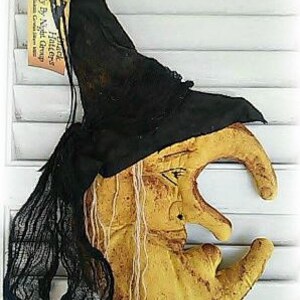 Puede incluir: Una luna creciente amarilla con cara de bruja y un sombrero de bruja negro. El sombrero tiene una etiqueta que dice "Witch Hat Fly By Night Creations".