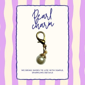 Könnte beinhalten: Ein goldfarbener Perlenanhänger mit einem Karabinerverschluss, der an einer kleinen Kette hängt. Der Anhänger befindet sich vor einem weißen Hintergrund mit den Worten "Pearl charm" in Schreibschrift. Der Hintergrund hat ein lila-weißes Streifenmuster.