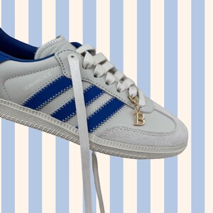 May include: White and blue trainer with blue stripes and the word "HUMANRACE" printed on the side. A gold charm with the letter "B" hangs from the laces. The shoe is set against a striped background of light blue and white.