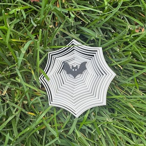 May include: A layered, white and black spiderweb decoration with a black bat silhouette in the center. The Halloween-themed decoration is set against a backdrop of green grass.