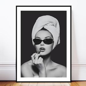 Puede incluir: Impresión enmarcada en blanco y negro de una mujer aplicándose lápiz labial. Lleva gafas de sol de ojo de gato y una toalla en la cabeza. La imagen tiene una estética vintage y glamurosa, con un enfoque en la belleza y el estilo.