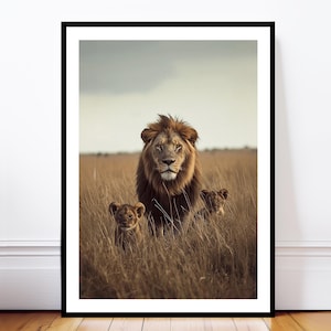 May include: A framed print depicting a male lion with a full mane and two lion cubs in long, dry grass. The artwork uses a natural colour palette of browns and tans, with a cloudy sky in the background. The image is framed in a black border.