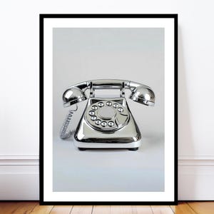 Pode incluir: Fotografia emoldurada de um telefone vintage com mostrador rotativo cromado. O telefone tem uma superfície brilhante e reflexiva e um cabo de auscultador enrolado. O mostrador apresenta números e o telefone está sobre um fundo branco.