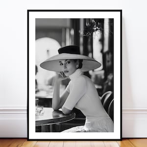 Puede incluir: Impresión enmarcada en blanco y negro de una mujer con un sombrero de ala ancha y un top blanco de cuello alto. Está sentada a una mesa, apoyando la barbilla en la mano. La imagen tiene una estética vintage clásica.