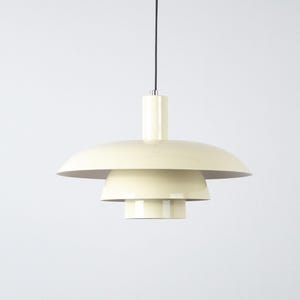 May include: Cream-colored pendant light with a tiered, saucer-like shade design. The light fixture hangs from a black cord and features a cylindrical top. The layered shades create a modern, minimalist aesthetic.
