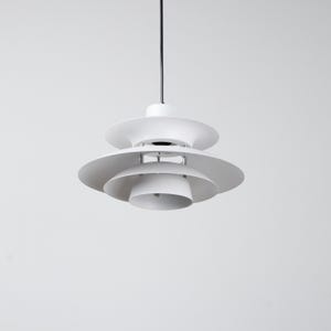 Op de afbeelding: Een witte, gelaagde hanglamp met een gelaagd, schotelachtig ontwerp. Het licht hangt aan een zwart snoer tegen een effen witte achtergrond. De armatuur heeft een moderne, minimalistische uitstraling.