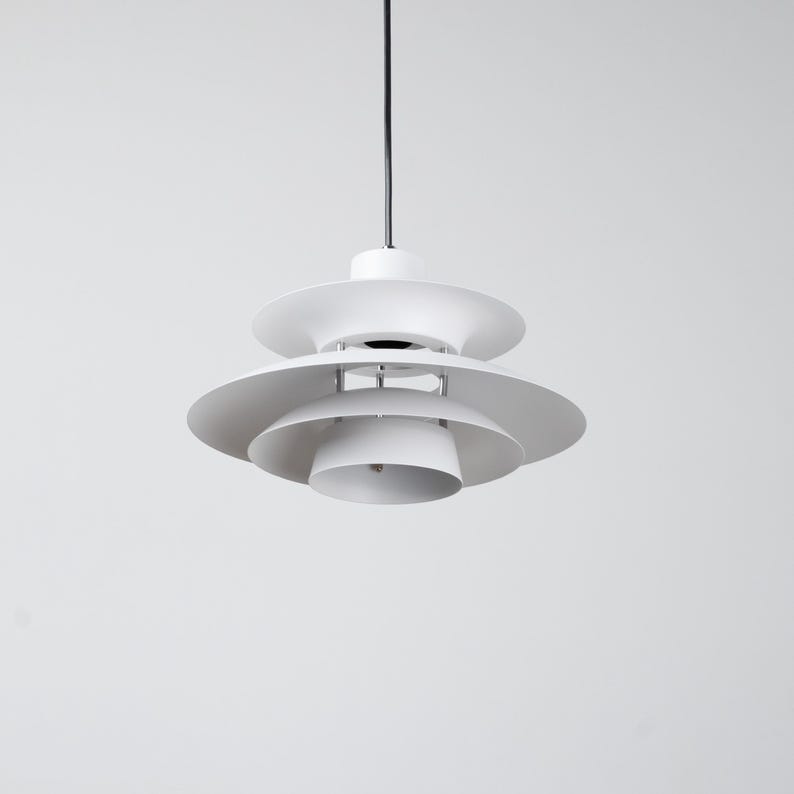 Op de afbeelding: Een witte, gelaagde hanglamp met een gelaagd, schotelachtig ontwerp. Het licht hangt aan een zwart snoer tegen een effen witte achtergrond. De armatuur heeft een moderne, minimalistische uitstraling.