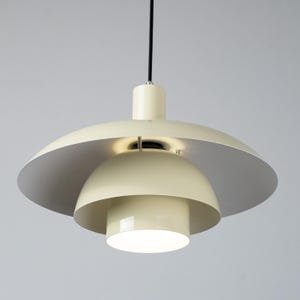 May include: Cream-colored pendant light fixture with a layered design. The light has a large, inverted saucer-shaped shade, a smaller dome-shaped shade, and a cylindrical base. The interior of the top shade is silver. A black cord suspends the light.