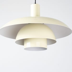 May include: Cream-colored, multi-tiered pendant light fixture. The light has a large, saucer-shaped shade, a rounded mid-section, and a cylindrical base. The interior of the shades is white. The light hangs from a black cord.