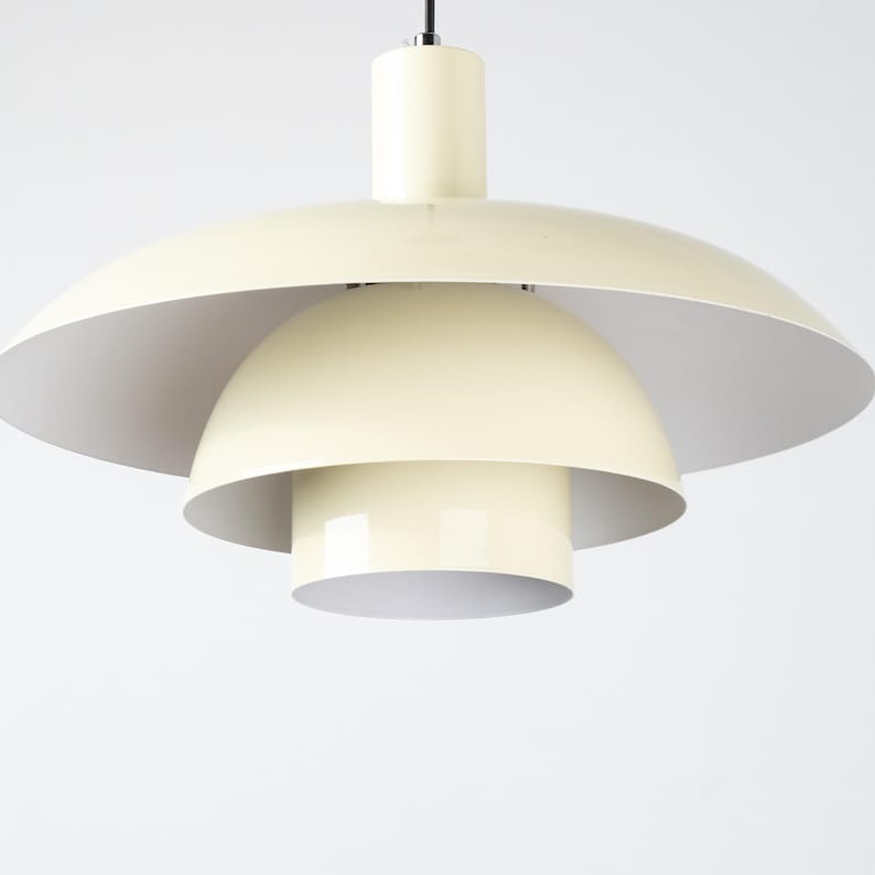 May include: Cream-colored, multi-tiered pendant light fixture. The light has a large, saucer-shaped shade, a rounded mid-section, and a cylindrical base. The interior of the shades is white. The light hangs from a black cord.