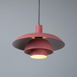 May include: A modern, hanging pendant light fixture in a soft coral color. The light has a layered design with a large, saucer-shaped top, a smaller dome, and a cylindrical bottom. The interior of the top and middle sections are silver.