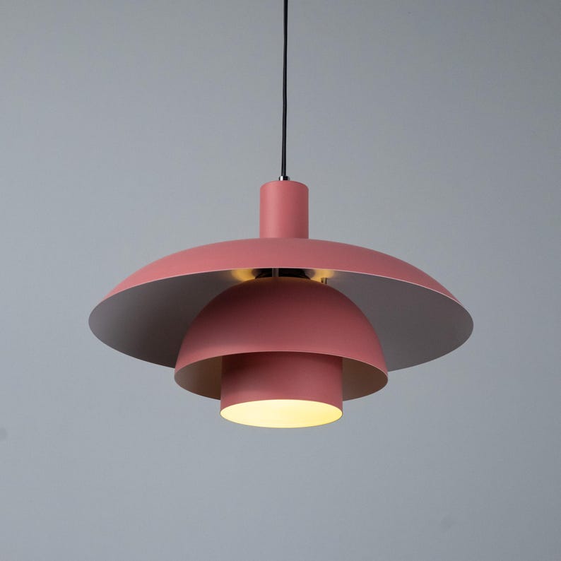 May include: A modern, hanging pendant light fixture in a soft coral color. The light has a layered design with a large, saucer-shaped top, a smaller dome, and a cylindrical bottom. The interior of the top and middle sections are silver.
