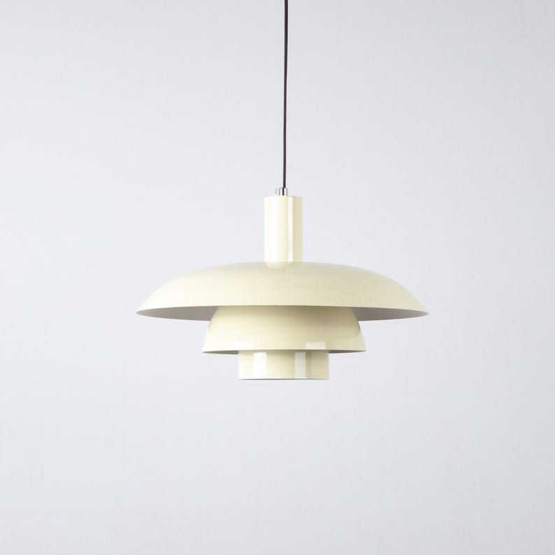 May include: Cream-colored pendant light with a tiered, saucer-like shade design. The light fixture hangs from a black cord and features a cylindrical top. The layered shades create a modern, minimalist aesthetic.