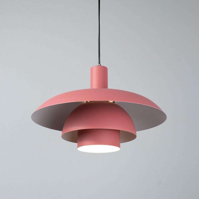 May include: A modern, layered pendant light fixture in a soft coral color. The light has a tiered design with a large, inverted saucer-shaped shade and a smaller, rounded shade below. The interior of the shades is a light, neutral color.