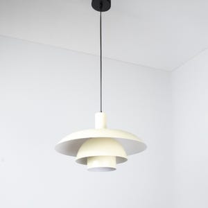 May include: Cream-colored pendant light fixture with a layered, saucer-like shade design. The light hangs from a black cord and ceiling mount. The fixture has a mid-century modern aesthetic.
