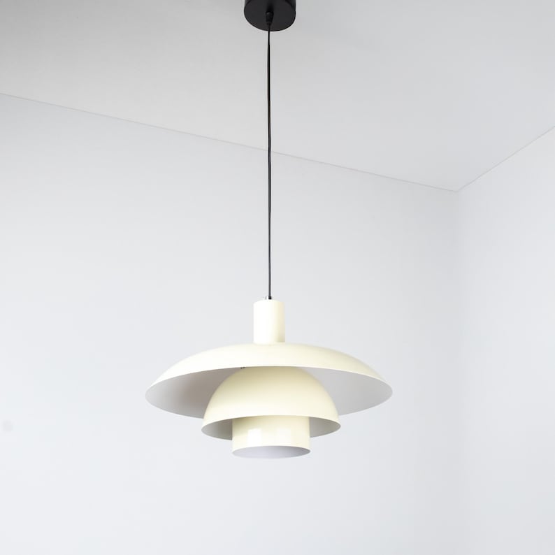 May include: Cream-colored pendant light fixture with a layered, saucer-like shade design. The light hangs from a black cord and ceiling mount. The fixture has a mid-century modern aesthetic.