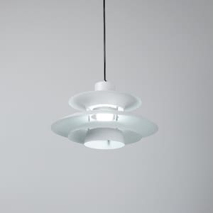 Op de afbeelding: Een witte, gelaagde hanglamp met een cirkelvormig ontwerp. De lamp heeft een reeks overlappende, schijfvormige tinten, waardoor een gelaagd effect ontstaat. De armatuur hangt aan een zwart snoer tegen een neutrale achtergrond.