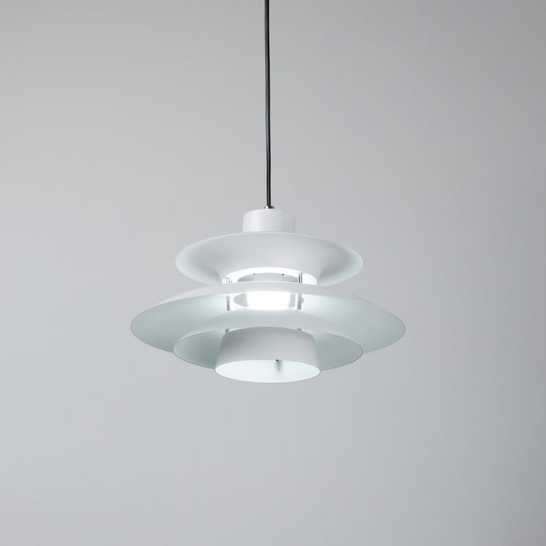 Op de afbeelding: Een witte, gelaagde hanglamp met een cirkelvormig ontwerp. De lamp heeft een reeks overlappende, schijfvormige tinten, waardoor een gelaagd effect ontstaat. De armatuur hangt aan een zwart snoer tegen een neutrale achtergrond.