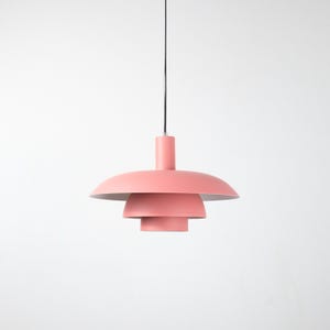 May include: A modern, tiered pendant light fixture in a soft coral color. The light has a cylindrical top and a series of descending, saucer-shaped shades. The fixture hangs from a black cord against a white background.