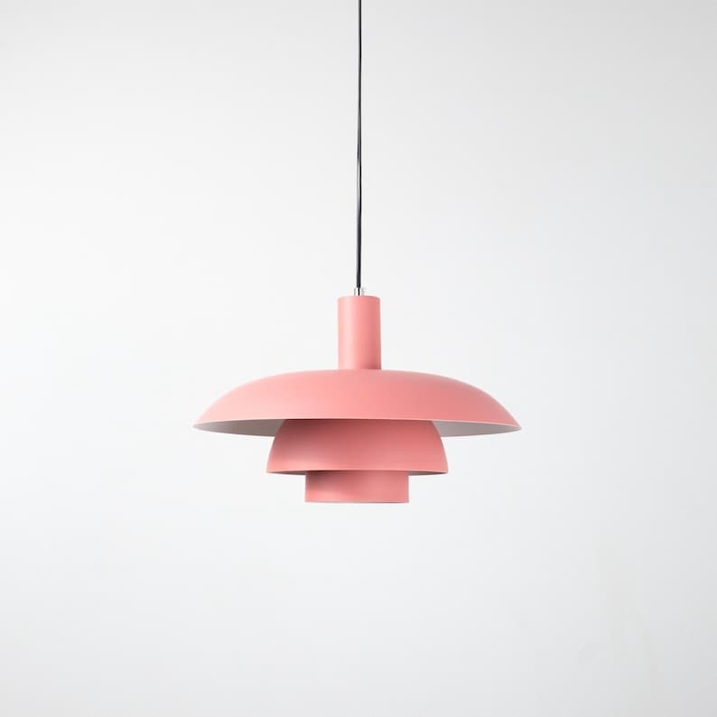 May include: A modern, tiered pendant light fixture in a soft coral color. The light has a cylindrical top and a series of descending, saucer-shaped shades. The fixture hangs from a black cord against a white background.