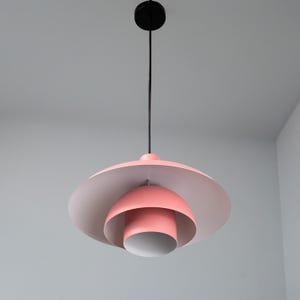 May include: A modern, pink and white pendant light fixture. The light has a black cord and ceiling mount. The light fixture has a layered design with a large, flat, circular shade and a smaller, rounded shade below.
