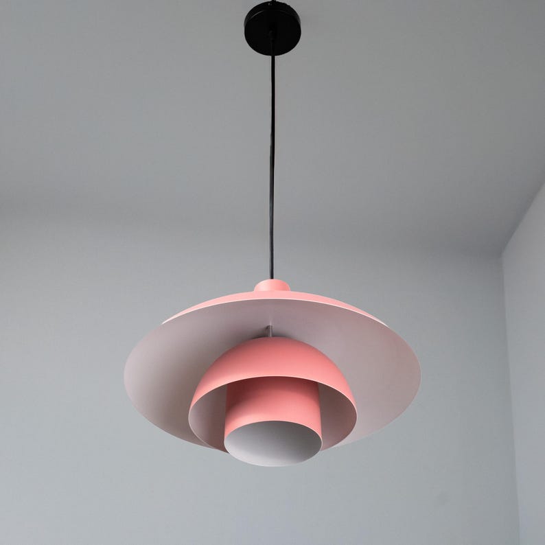 May include: A modern, pink and white pendant light fixture. The light has a black cord and ceiling mount. The light fixture has a layered design with a large, flat, circular shade and a smaller, rounded shade below.