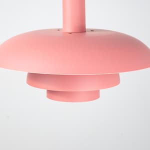 May include: A modern, tiered pendant light fixture in a soft coral color. The light has a circular, layered design with a central stem. The light is suspended from the ceiling, creating a minimalist aesthetic.