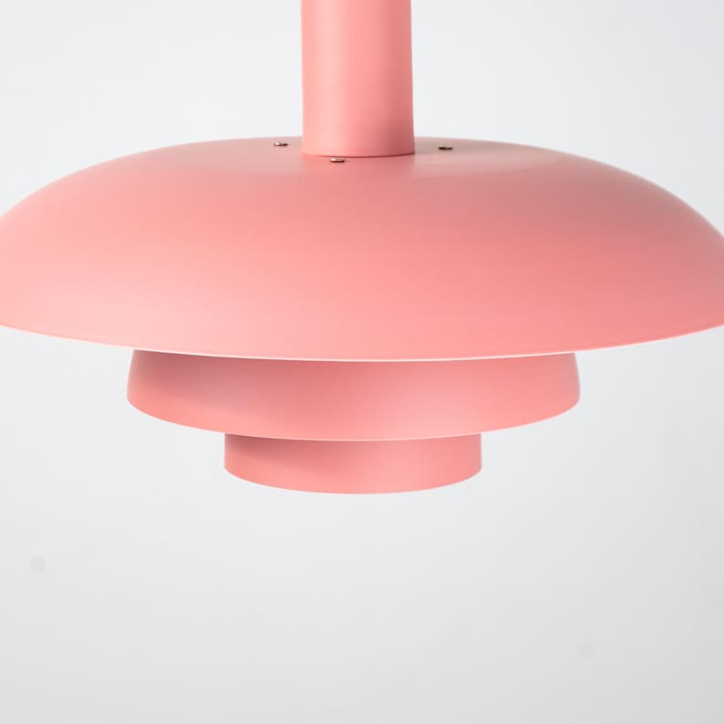 May include: A modern, tiered pendant light fixture in a soft coral color. The light has a circular, layered design with a central stem. The light is suspended from the ceiling, creating a minimalist aesthetic.