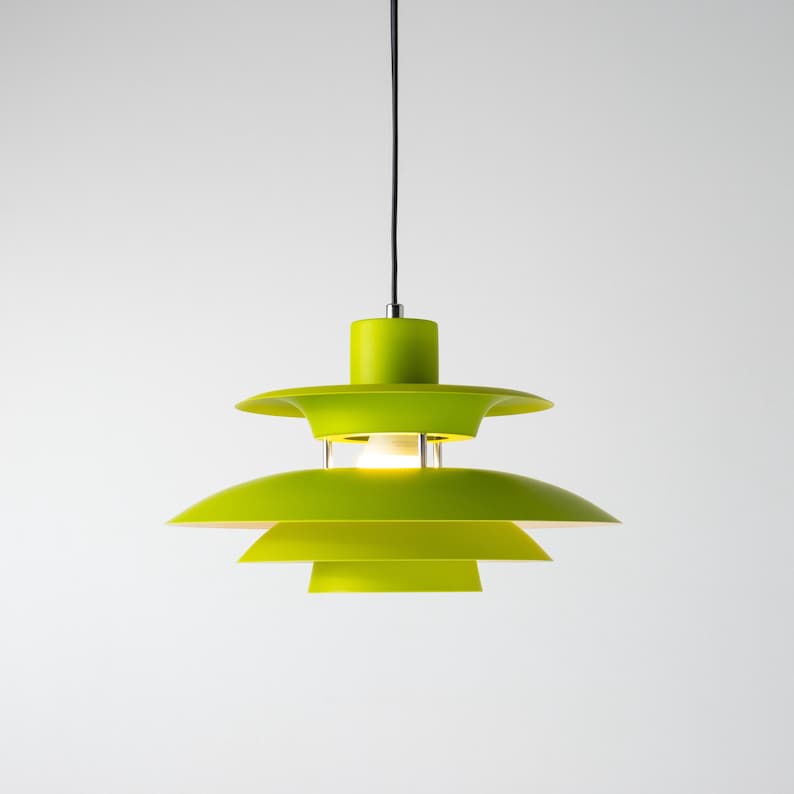 Op de afbeelding: Een limoengroene hanglamp met een gelaagd, schotelvormig schaduwontwerp. Het licht hangt aan een zwart snoer en heeft een cilindrische bovenkant. De armatuur hangt tegen een witte achtergrond.
