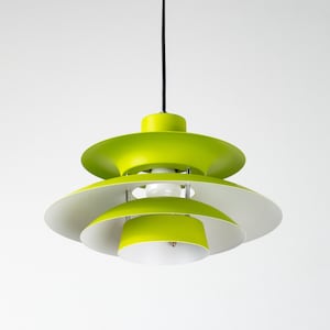 Op de afbeelding: Een fel limoengroene en witte hanglamp. De lamp heeft een gelaagd, schotelachtig ontwerp, met een zwart snoer. De binnenkant is wit, wat een retro esthetiek cre&euml;ert. De lamp hangt tegen een witte achtergrond.