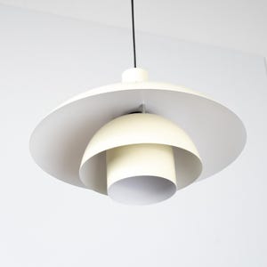 May include: A modern, cream-colored pendant light fixture. The design features a large, flat, circular shade above a layered, dome-shaped structure. A cylindrical element hangs below, all suspended from a black cord.