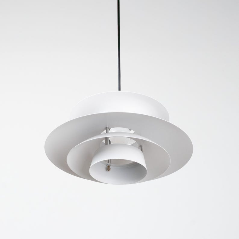 Op de afbeelding: Een witte, gelaagde hanglamp met een cirkelvormig ontwerp. De lamp heeft meerdere niveaus van gebogen, schijfvormige kappen, opgehangen aan een zwart snoer. De armatuur is een modern, minimalistisch ontwerp.