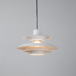 Op de afbeelding: Een witte, gelaagde hanglamp met een cirkelvormig ontwerp. De lamp heeft overlappende, schotelvormige kappen, opgehangen aan een zwart snoer. De armatuur straalt een warm licht uit, geschikt voor moderne interieurs. De diameter is ongeveer 30 cm.