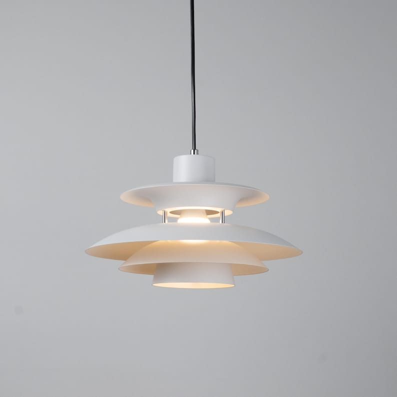 Op de afbeelding: Een witte, gelaagde hanglamp met een cirkelvormig ontwerp. De lamp heeft overlappende, schotelvormige kappen, opgehangen aan een zwart snoer. De armatuur straalt een warm licht uit, geschikt voor moderne interieurs. De diameter is ongeveer 30 cm.