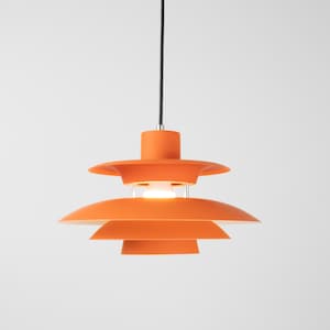 Op de afbeelding: Oranje hanglamp met een gelaagd, trapsgewijs ontwerp. De lamp heeft een cilindrische bovenkant en een reeks afdalende, schotelvormige kappen. Een zwart snoer hangt het licht op tegen een witte achtergrond.