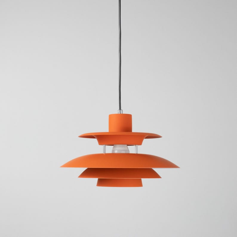 Op de afbeelding: Oranje hanglamp met een gelaagd ontwerp. De lamp hangt aan een zwart snoer en heeft een cilindrische bovenkant. De gloeilamp is zichtbaar in het midden van de lamp.