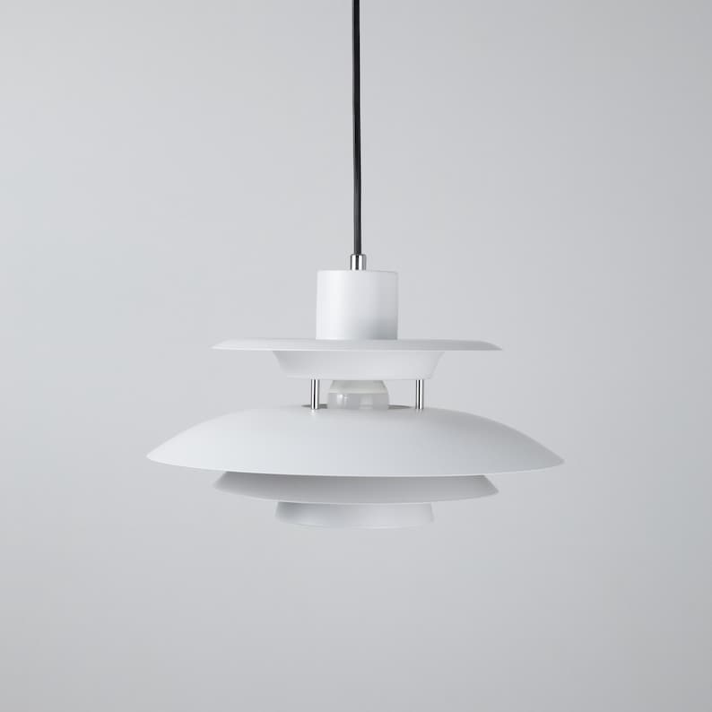 Op de afbeelding: Een witte, moderne hanglamp met een meerlaags kapontwerp. De lamp heeft een cirkelvormige bovenkant, een middelste laag en een onderste laag, allemaal in het wit. De lamp hangt aan een zwart snoer en is geschikt voor binnenverlichting.