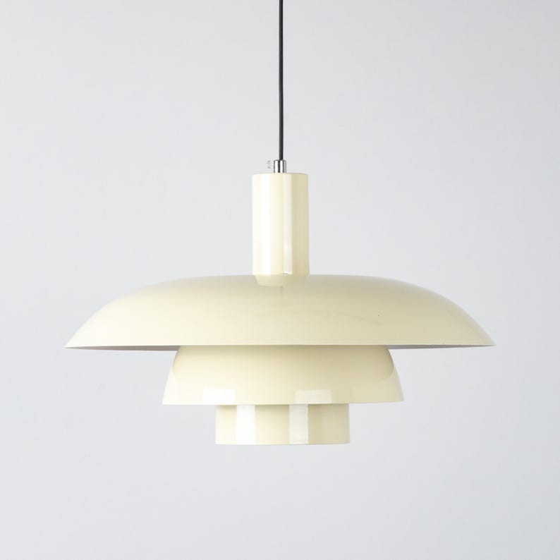 May include: Cream-colored pendant light with a tiered, saucer-like shade. The light fixture has a cylindrical top and a black cord. The design is minimalist and modern, suitable for various interior styles.