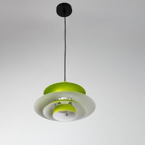 Op de afbeelding: Een moderne hanglamp met een gelaagd ontwerp. De lamp heeft een zwarte plafondbevestiging, een zwart snoer en overlappende, cirkelvormige kappen in wit, groen en wit. De lamp hangt tegen een effen witte achtergrond.