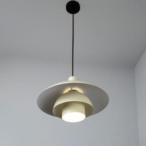 May include: A modern pendant light fixture with a circular, layered design. The light features a cream-colored, bowl-shaped shade with a gray outer ring. A black cord and ceiling mount suspend the light, which is illuminated from within.