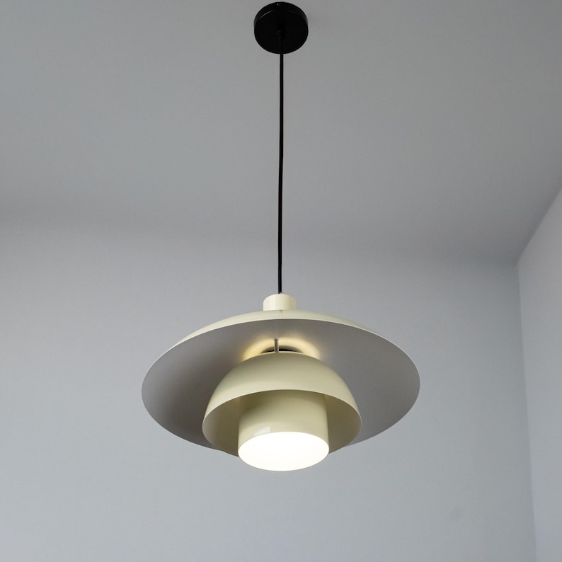 May include: A modern pendant light fixture with a circular, layered design. The light features a cream-colored, bowl-shaped shade with a gray outer ring. A black cord and ceiling mount suspend the light, which is illuminated from within.