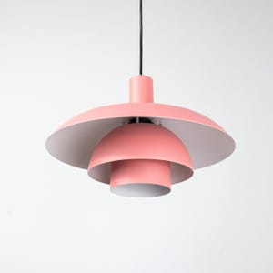 May include: A modern, tiered pendant light fixture in a soft coral pink. The light has a layered design with a large, saucer-shaped shade and a smaller, rounded shade below. The interior of the shades is white, and the fixture hangs from a black cord.
