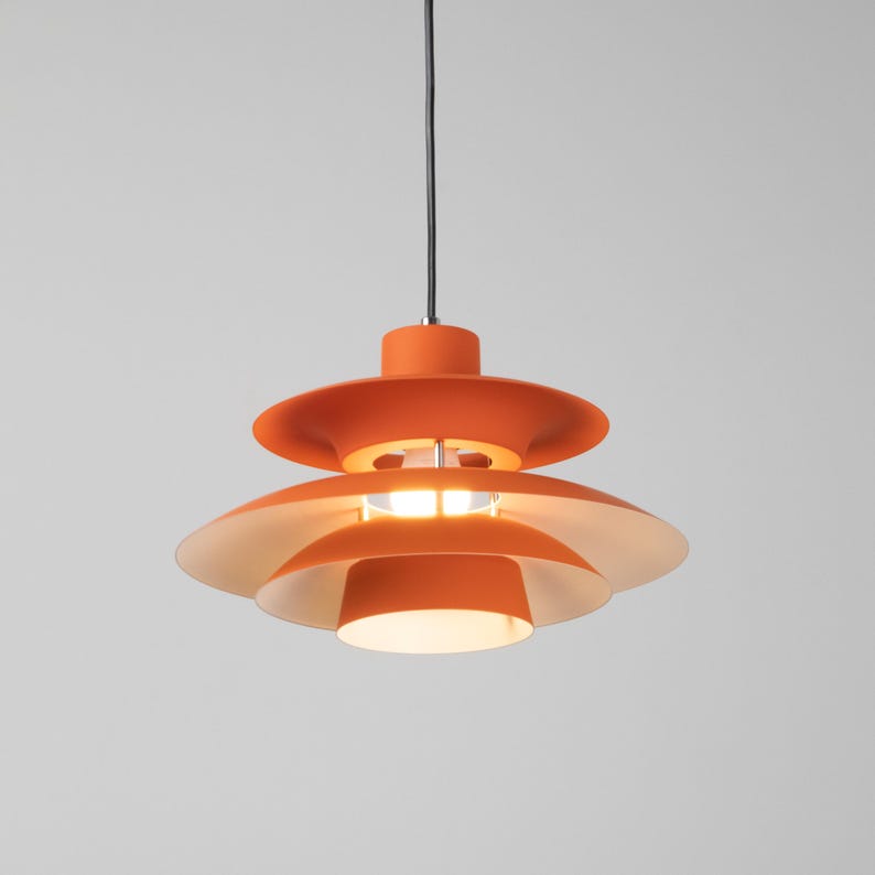 Op de afbeelding: Oranje hanglamp met een gelaagd, schijfvormig ontwerp. De lamp heeft een centrale lamp en een zwart snoer. De armatuur hangt tegen een neutrale achtergrond en toont zijn moderne esthetiek. Diameter: ongeveer 30 cm.