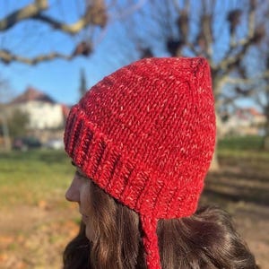 May include: A vibrant red knit beanie featuring a textured ribbed cuff and two long braided tassels hanging from the sides. The yarn displays a subtle heathered effect with flecks of lighter red and white throughout the thick, warm construction.