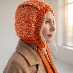 Könnte beinhalten: Eine leuchtend orangefarbene Strickhaube mit einer strukturierten, flauschigen Oberfläche, die mit kleinen, reflektierenden goldenen Pailletten verziert ist. Die Kopfbedeckung hat einen gerippten Strickrand und zwei lange, passende orangefarbene Bänder.